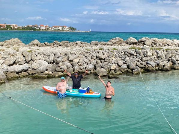 Three people cheering in shallow turquoise water of a tropical coastal cove, using a colorful paddleboard as a floating drinks station near a rocky breakwater with resort homes on the distant shoreline.