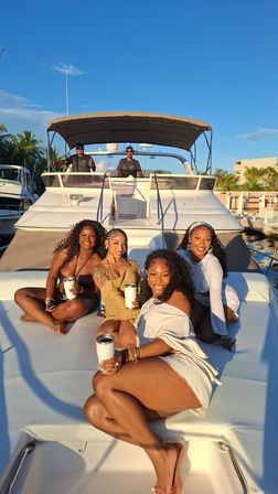 Four women laughing and lounging on the bow of a white yacht at a sunny tropical marina, holding travel tumblers and enjoying golden-hour light