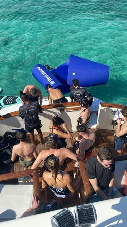 Aerial view of a group in snorkel gear on a wooden boat deck next to a bright blue floating water ramp in crystal-clear turquoise sea