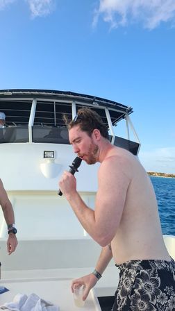 Shirtless man with sunglasses on his head singing into a microphone and holding a drink on a boat deck, clear blue ocean and sky in the background — sunny tropical karaoke vacation scene