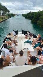 Passengers relaxing on a leisure charter boat cruising a tropical mangrove-lined channel toward sailboats, with palm trees and a cloudy sky