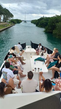 Passengers relaxing on a leisure charter boat cruising a tropical mangrove-lined channel toward sailboats, with palm trees and a cloudy sky