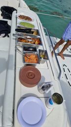 Onboard boat buffet with taco-style toppings: trays of pico de gallo, guacamole, chips, grilled chicken, black beans and rice arranged on a white yacht rail beside turquoise water, with stacked purple plates, utensils and a drink nearby.
