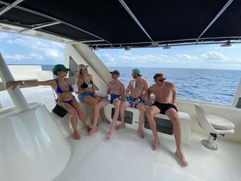 Five friends relaxing on a yacht deck under a shaded canopy, wearing swimsuits and caps and holding drinks with the open ocean and blue sky on a sunny day.