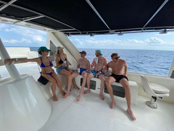 Five friends relaxing on a yacht deck under a shaded canopy, wearing swimsuits and caps and holding drinks with the open ocean and blue sky on a sunny day.
