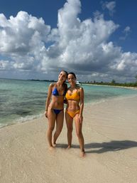 Two friends in blue and yellow bikinis standing in shallow turquoise water on a white-sand tropical beach, with an anchored boat in the distance and dramatic puffy clouds overhead.