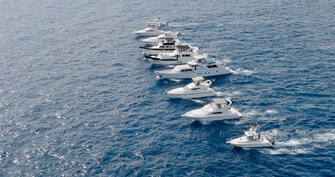 Aerial view of a diagonal fleet of white luxury yachts cruising across deep blue open ocean, each leaving foamy wakes in clear water.