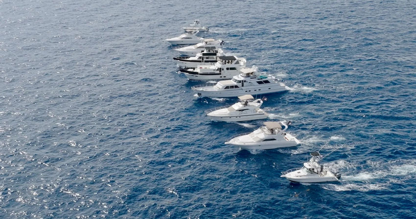 Aerial view of a diagonal fleet of white luxury yachts cruising across deep blue open ocean, each leaving foamy wakes in clear water.