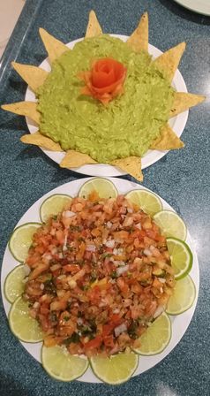 Overhead shot of Mexican-style appetizers: a white plate of creamy guacamole topped with a tomato rose and tortilla chips arranged like sun rays, and a second plate of fresh pico de gallo salsa surrounded by lime slices on a kitchen countertop.
