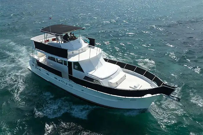 Aerial view of a white luxury motor yacht with black trim cruising on clear turquoise ocean under a sunny sky, passengers on the shaded flybridge and a gentle wake trailing behind.