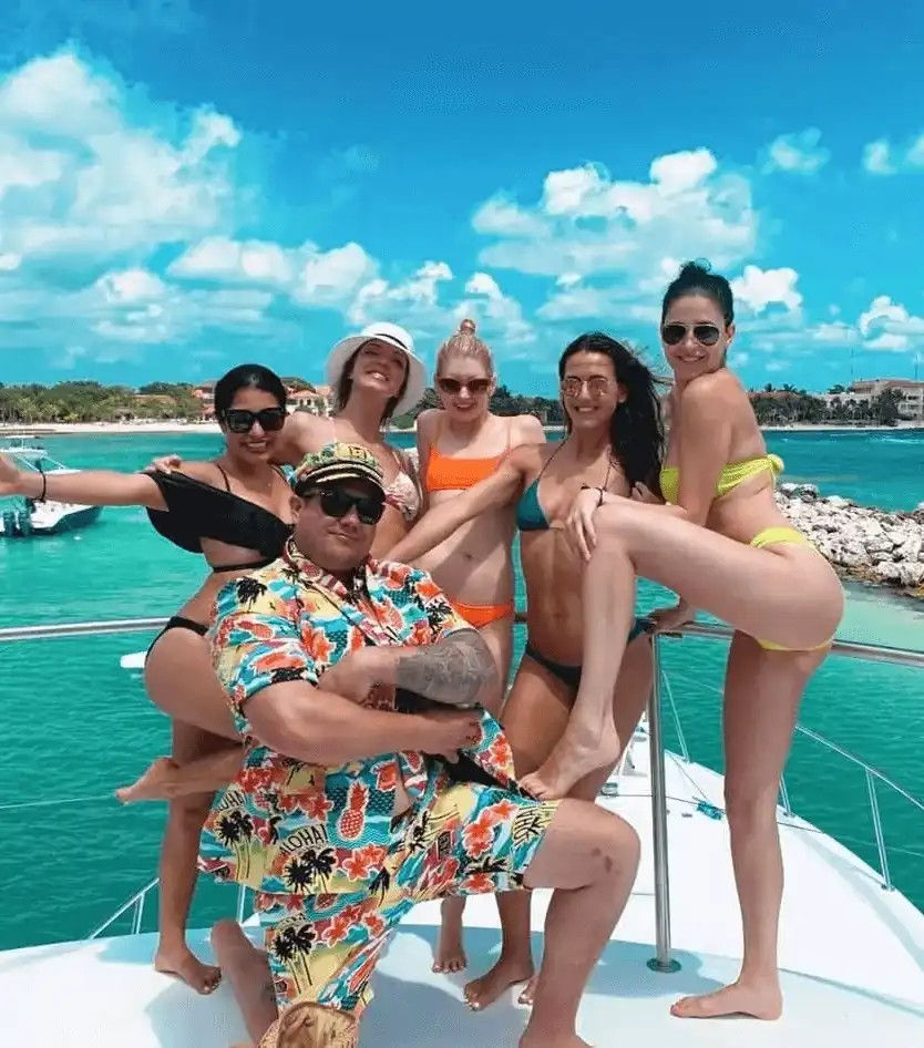 Festive yacht scene with a man in a colorful Hawaiian shirt and captain hat posing with five women in bikinis on a sunlit deck, turquoise water and tropical coastline under a bright blue sky.