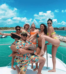 Festive yacht scene with a man in a colorful Hawaiian shirt and captain hat posing with five women in bikinis on a sunlit deck, turquoise water and tropical coastline under a bright blue sky.