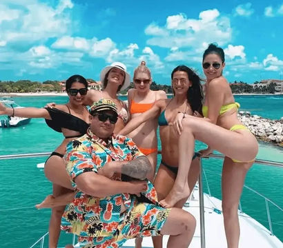 Festive yacht scene with a man in a colorful Hawaiian shirt and captain hat posing with five women in bikinis on a sunlit deck, turquoise water and tropical coastline under a bright blue sky.