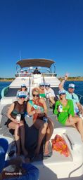 Group of friends lounging on the bow of a white yacht under a clear blue sky, wearing matching caps and holding drinks; central woman in a sash and floral headband with others smiling and flashing peace signs while cruising coastal waters near a rocky shoreline.