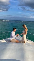 Romantic boat proposal on a turquoise sea — a person kneeling on the bow offering a ring to another, with blue sky and scattered clouds overhead.