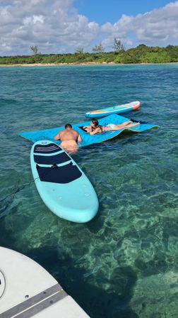Two people lounging on a blue floating mat next to turquoise paddleboards in crystal-clear tropical water by a sandy shoreline under a sunny, partly cloudy sky