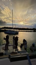 Silhouetted people on a yacht deck snapping photos of an anchored catamaran as golden sunset rays light up a calm coastal harbor.