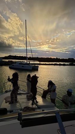 Silhouetted people on a yacht deck snapping photos of an anchored catamaran as golden sunset rays light up a calm coastal harbor.