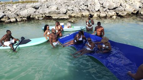 Sunny beach scene: a group enjoying drinks on paddleboards and a large blue floating mat in clear turquoise coastal water near a rocky shoreline.