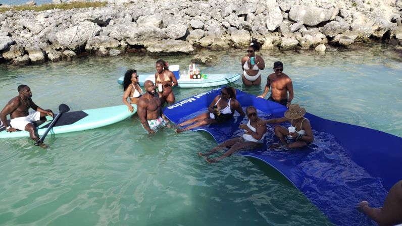 Sunny beach scene: a group enjoying drinks on paddleboards and a large blue floating mat in clear turquoise coastal water near a rocky shoreline.