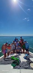 Cheerful group on the bow of a boat in clear turquoise ocean under a bright sunny sky, many wearing matching caps and swimwear, one wearing a sash, with towels and beach gear scattered on the deck.