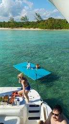 Sunny tropical boating scene: kids playing on a bright blue floating mat tied to a boat in clear turquoise water, an adult on the swim platform with snacks on deck and a sandy, tree-lined shoreline in the background.