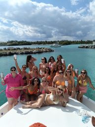 Group of women on a yacht enjoying a bikini boat party in turquoise tropical waters near a rocky, palm-lined island under a sunny sky with clouds