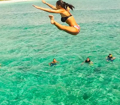 Woman leaping into crystal-clear turquoise ocean near a tropical island shoreline with snorkelers floating below