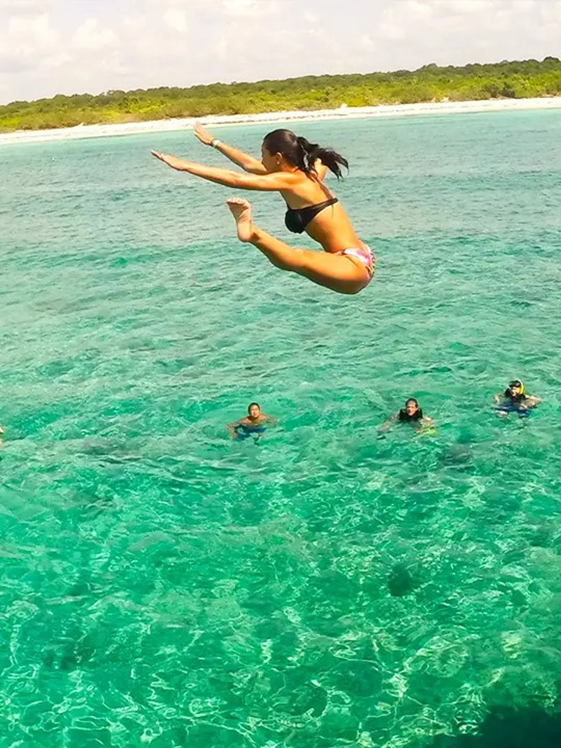 Woman leaping into crystal-clear turquoise ocean near a tropical island shoreline with snorkelers floating below