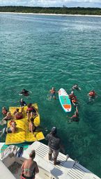 View from a boat's swim platform of a lively group in clear turquoise water—swimmers and life-jacketed guests clustered around a yellow floating mat and a colorful paddleboard, with a sandy tree-lined shoreline beyond.