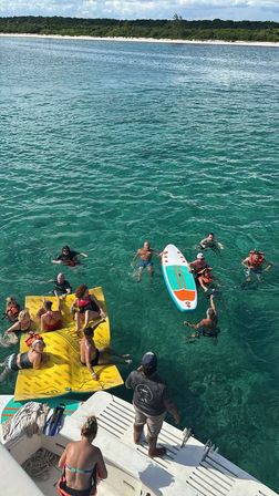 View from a boat's swim platform of a lively group in clear turquoise water—swimmers and life-jacketed guests clustered around a yellow floating mat and a colorful paddleboard, with a sandy tree-lined shoreline beyond.
