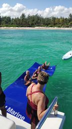 Group of women lounging on a blue floating mat tied to a boat in clear turquoise water off a palm‑lined tropical beach under a sunny sky