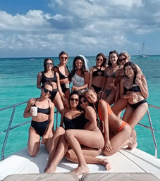 Group of women in bikinis posing on the bow of a yacht over clear turquoise tropical water under a partly cloudy sky, holding drinks and smiling for a lively boat party — one wearing a bridal veil.