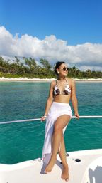 Woman in cow-print bikini and white sarong sitting on a boat railing over turquoise tropical water with a palm-lined sandy beach and blue sky with clouds in the background