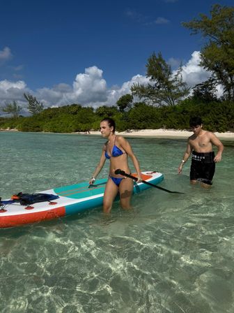 Two young adults wading with a colorful stand-up paddleboard in clear turquoise shallow water off a white-sand tropical beach lined with green trees under a blue sky with puffy clouds