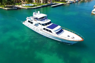 Aerial view of a sleek white luxury yacht gliding through crystal-turquoise canal past palm-lined waterfront homes and private docks.