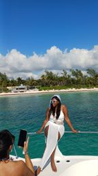 Woman in white dress with veil and heart sunglasses posing on a boat over turquoise tropical water, palm-lined sandy beach and blue sky with fluffy clouds