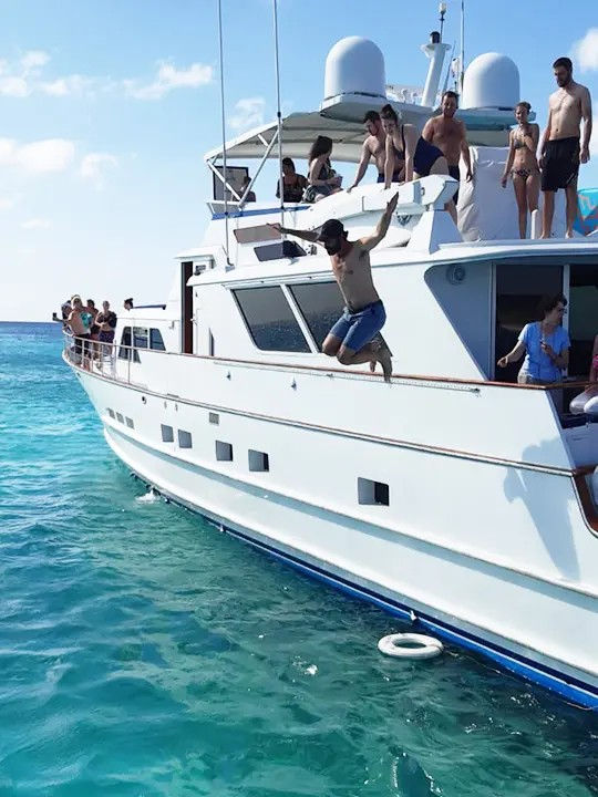 Group of people on a white yacht with a man mid-jump into clear turquoise ocean on a sunny boat-party vacation