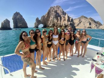 Group of women in bikinis posing on a boat with the natural rock arch El Arco and turquoise waters of Cabo San Lucas, Mexico on a sunny day