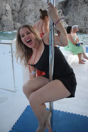Woman in a black dress playfully posing on a dance pole aboard a boat near rocky coastal cliffs, smiling during a seaside boat party with ocean in the background.