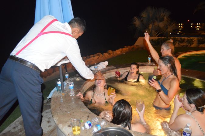 Nighttime outdoor hot tub party with friends laughing and holding cocktails while a man in a white shirt and pink suspenders leans over the pool edge to pour a drink into a woman’s mouth; pool deck, water bottles, and lights in the background.