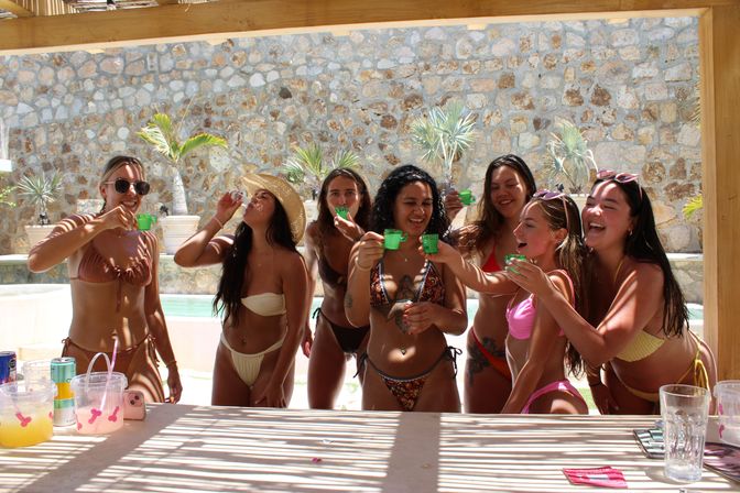Seven women in colorful bikinis laughing and clinking green shot cups at a sunny tropical poolside bar with a stone wall and potted palms.