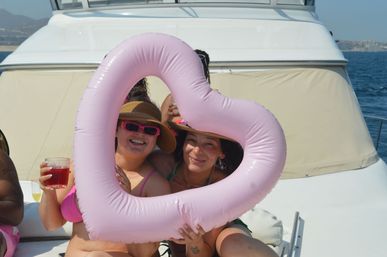 Two friends on a sunny yacht holding a pink heart-shaped inflatable float, wearing sunhats and sunglasses, one holding a red drink with ocean and coastline in the background.