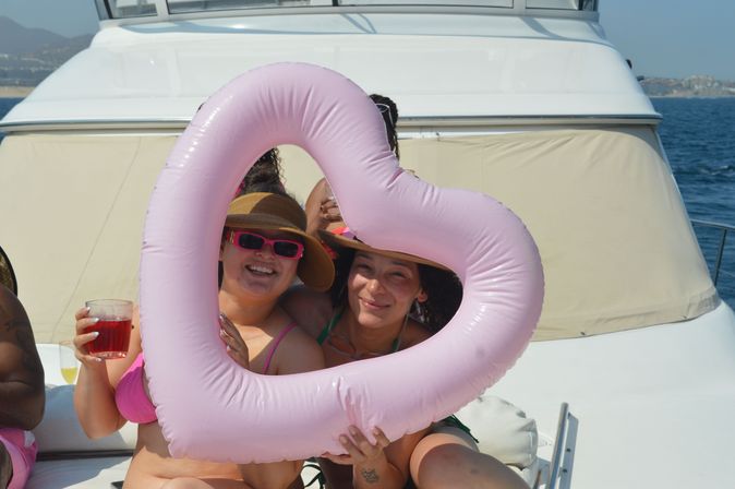Two friends on a sunny yacht holding a pink heart-shaped inflatable float, wearing sunhats and sunglasses, one holding a red drink with ocean and coastline in the background.