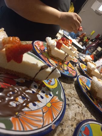 Strawberry-topped cheesecake slices with chocolate drizzle on colorful ceramic plates lined up on a granite kitchen counter, a hand placing fresh berries.