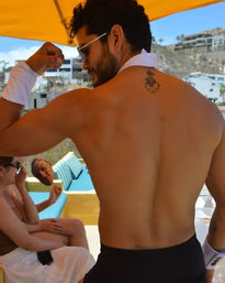 Shirtless man flexing back and bicep under a yellow beach umbrella at a sunny seaside resort poolside cabana, wearing sunglasses and white wristbands with a small upper-back tattoo and people lounging on blue cushions in the background.