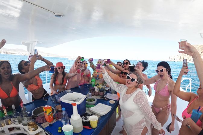 Group of women in colorful bikinis cheering with shot glasses at a lively boat party on a yacht over clear blue ocean and sunny coastal backdrop