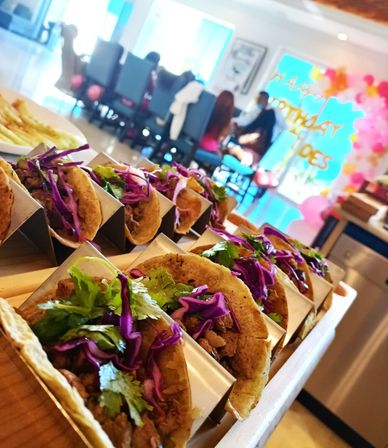 Close-up of a catering spread: row of soft tacos with shredded beef, purple cabbage and cilantro in metal holders on a wooden board at a colorful indoor birthday party with pink balloon arch in the background.