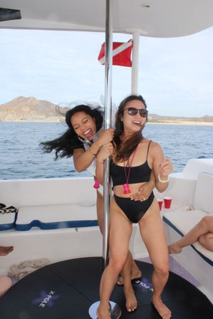Two women laughing and holding a chrome dance pole on a sunny party boat, wearing swimsuits with ocean and rocky coastline in the background.