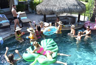 Sunny backyard pool party with a group of friends in colorful bikinis and sun hats, people enjoying drinks on a green leaf float and clear inflatable near a thatched cabana and poolside lounge chairs.